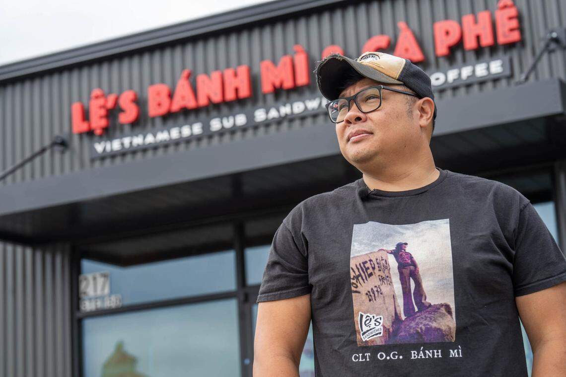 The owner of Le’s Sandwiches stands in front of the restaurant’s sign wearing a trucker hat, glasses, and a black t-shirt.