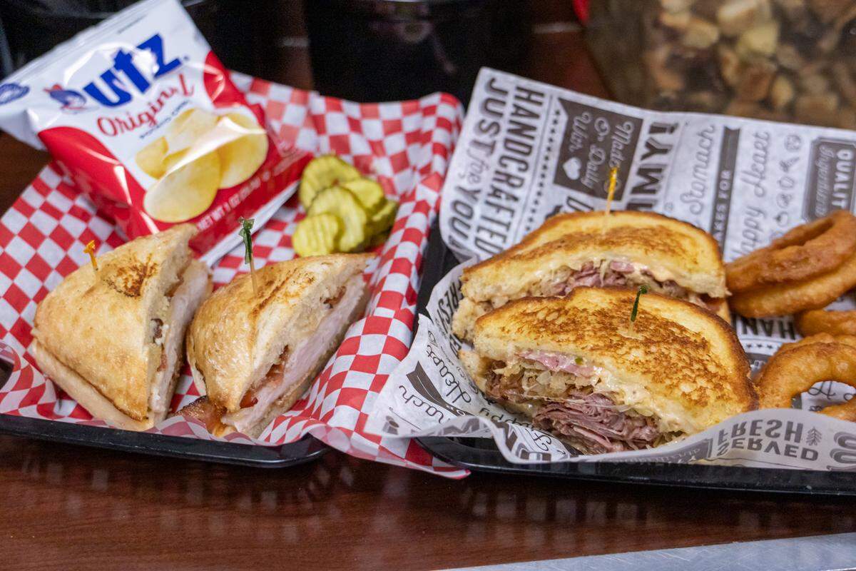 A top-down shot of two black trays filled with deli favorites. The tray on the left features a toasted club sandwich on a sourdough-style roll, served with crinkle-cut pickles and a small bag of Utz Original potato chips in a red and white checkered liner. The tray on the right holds a classic Reuben sandwich, sliced in half to show the corned beef, sauerkraut, and melted cheese, served with a side of golden-brown onion rings on branded parchment paper.