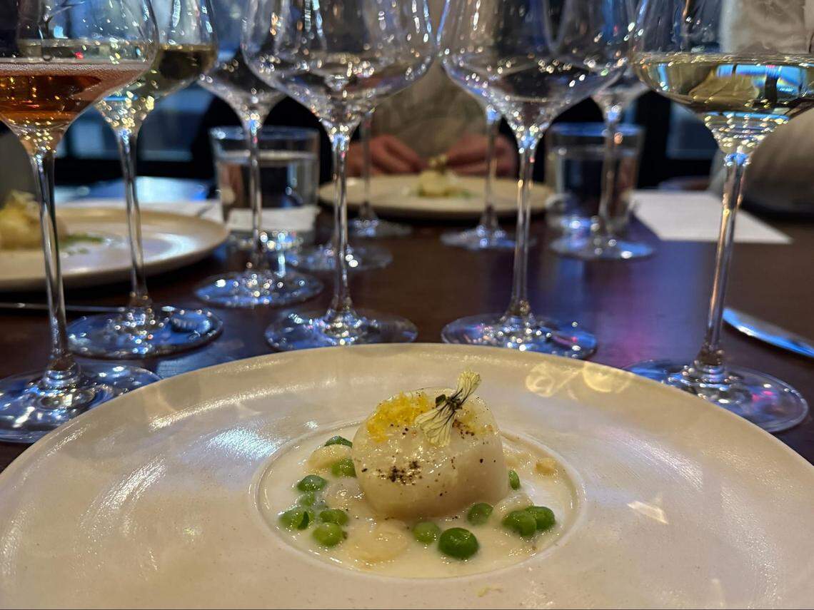 A close-up, low-angle shot of a meticulously plated scallop or quenelle dish served in a shallow pool of creamy white sauce with green peas on a wide white plate. A garnish of zest and a small white flower is placed on top of the food. In the background, several wine glasses filled with white and rosé wine are lined up on a dark wood table, with other blurred plates and diners visible further back.
