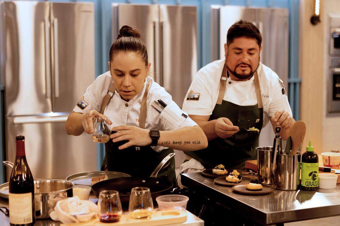 Two chefs in white coats and dark aprons work intently in a professional kitchen. The chef in the foreground is carefully pouring liquid from a glass, while the chef behind him plates small appetizers on a stainless steel counter.