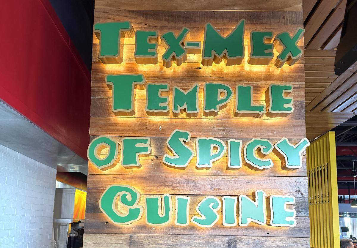 A close-up of a backlit decorative sign mounted on a rustic wood-plank wall. The sign features large, stylized green 3D letters that read “Tex-Mex Temple of Spicy Cuisine.” A warm orange glow emanates from behind each word, highlighting the uneven, playful font.