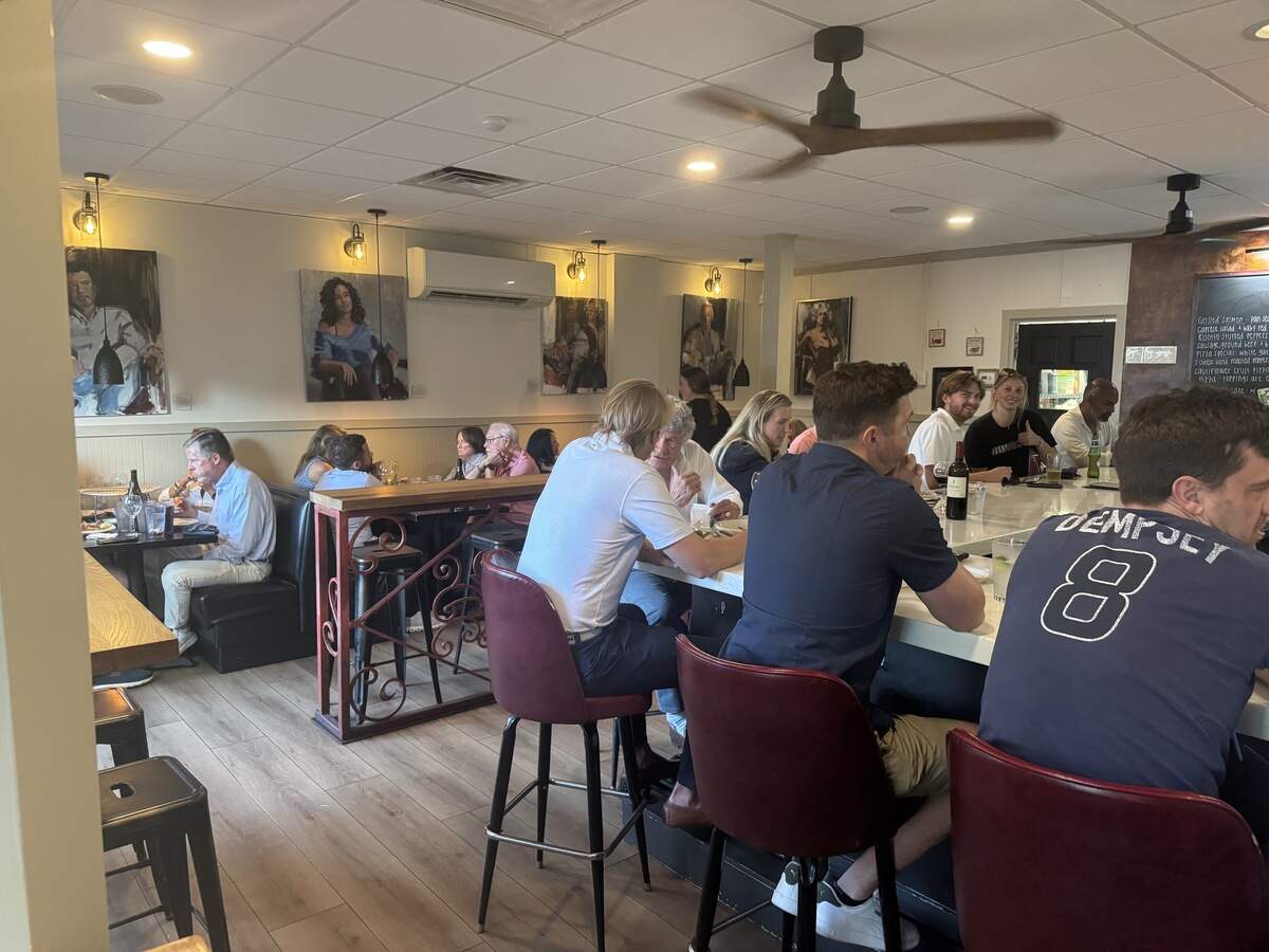 The bustling interior of a casual dining restaurant. Patrons are seated along a long, white-countertop bar on dark red and black barstools, while others sit in booths against the wall. The space features light-colored walls adorned with painted portraits, warm pendant lighting, wood flooring, and a ceiling fan in motion. The atmosphere appears lively and conversational.