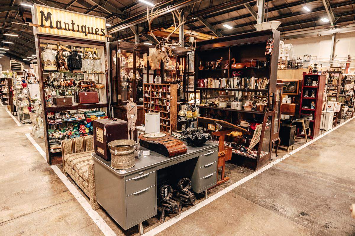 An antique mall booth is showcased, with a prominent lit sign that reads “Mantiques.” In the foreground, a vintage grey metal desk and a patterned bench are displayed. The back of the booth is filled with dark wood shelving holding books, pottery, and glassware, and a large taxidermy deer head is mounted on a cabinet.