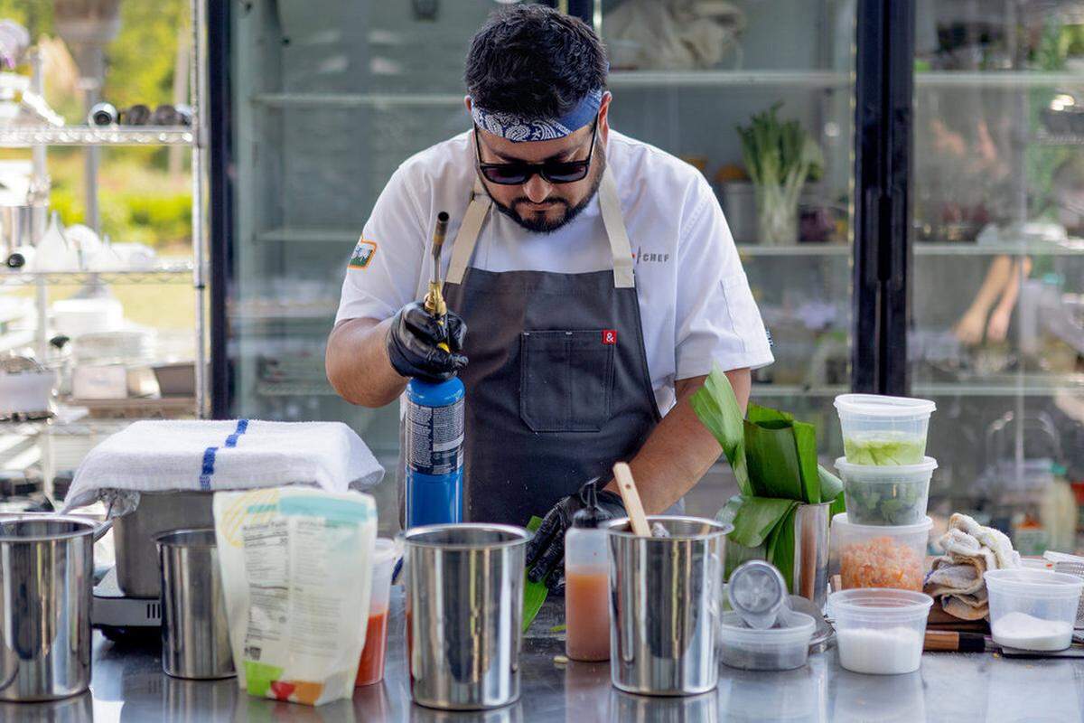 A focused, professional shot of a chef at an outdoor cooking station. The chef, wearing a white coat, a dark grey apron, a blue patterned bandana, and sunglasses, uses a blue blowtorch to carefully char green leaves on a stainless steel prep table.