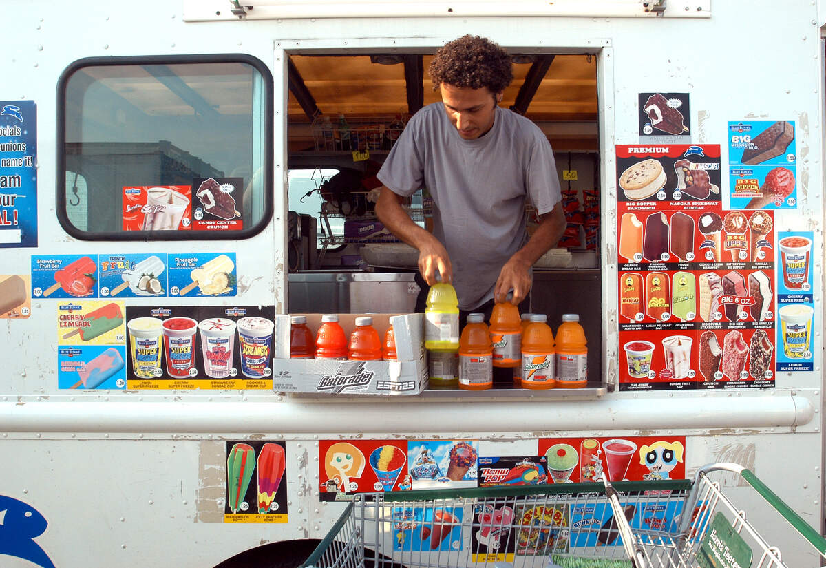 An ice cream truck operator arranges bottles of drinks on the service counter. The side of the white truck is covered in colorful menu decals displaying various frozen treats.