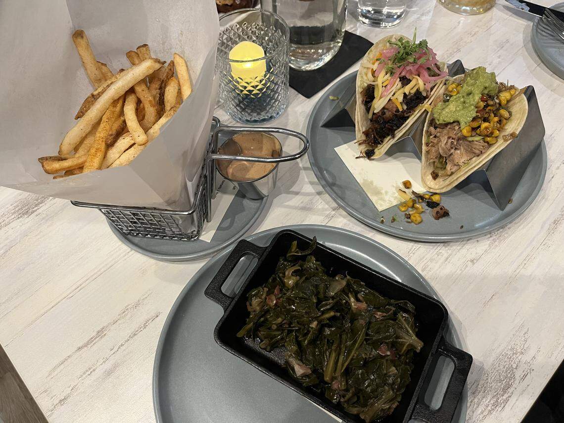 A high-angle shot displays three dishes arranged on a light-colored, wood-grain table: a black rectangular dish of collard greens in the foreground, a wire basket of french fries with a dipping sauce to the upper left, and a plate holding two tacos in a stand to the upper right.
