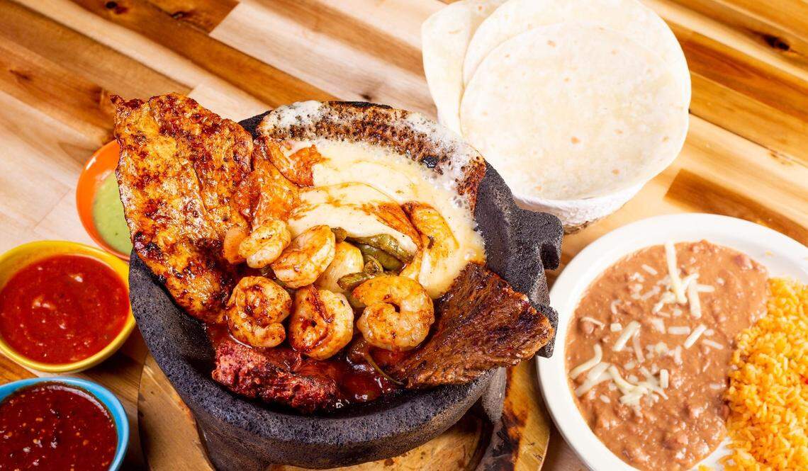 A top-down shot of a large and vibrant Mexican meal served on a wooden table. In the center, a sizzling black volcanic rock bowl (molcajete) holds a bubbling mix of melted white cheese, grilled shrimp, a chicken breast, and a piece of carne asada. To the side, a basket holds a stack of flour tortillas, and a white plate is filled with Spanish rice and refried beans topped with shredded cheese. Three small colorful bowls of salsa and guacamole are also arranged on the table.