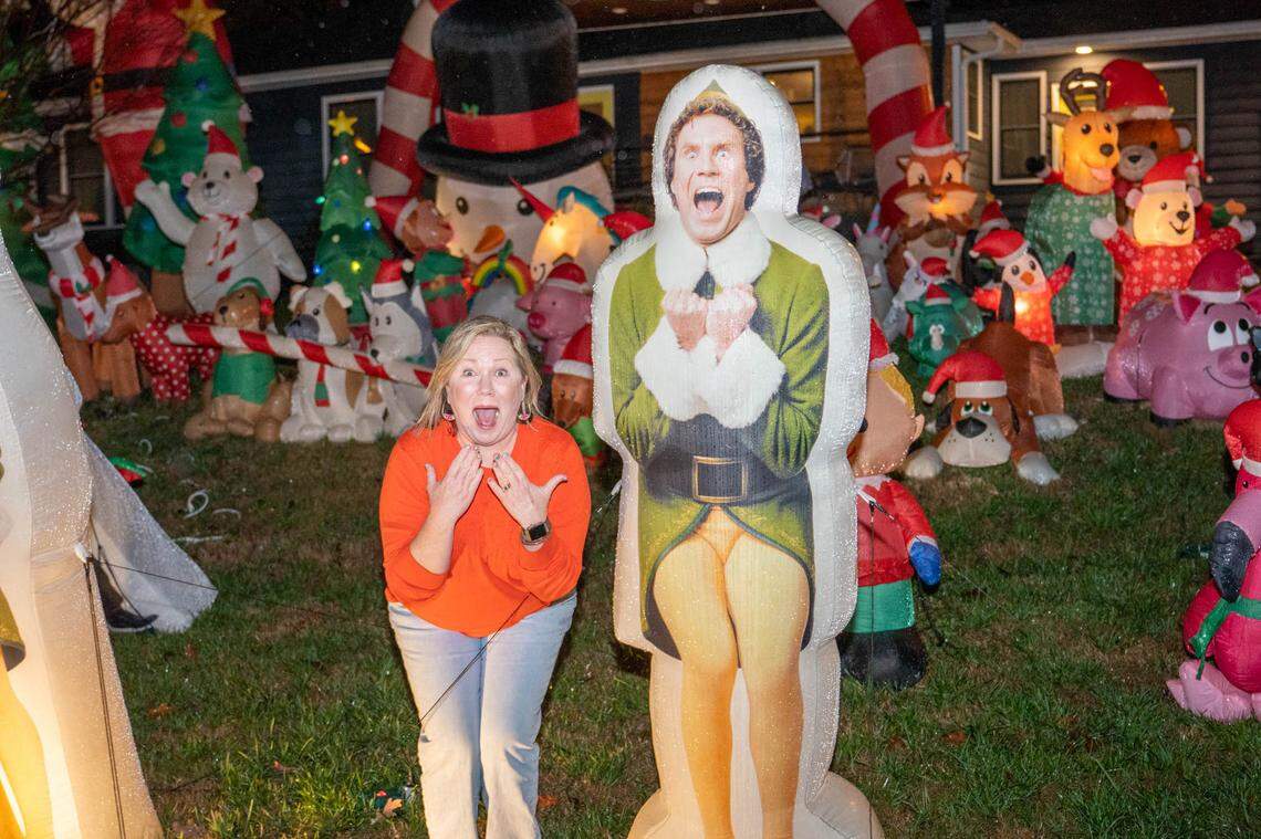 A playful nighttime photo showing a woman mimicking the excited, wide-eyed expression of a large inflatable cutout of Buddy the Elf from the movie Elf. Behind them, the lawn is completely filled with a large collection of colorful, inflatable Christmas characters, including Santa, snowmen, and animals.
