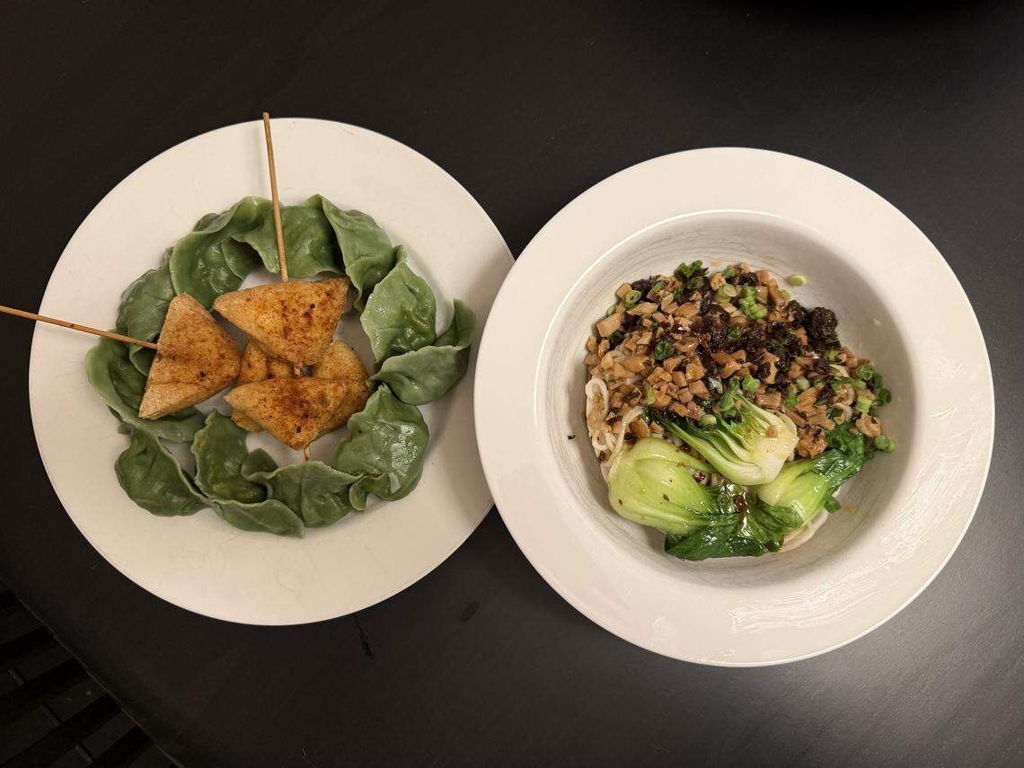 An overhead shot of two white plates of Asian cuisine on a dark surface. The left plate holds green dumplings arranged around three pieces of fried or pan-seared triangle-shaped food on wooden skewers. The right plate contains noodles topped with ground meat, a dark sauce, chopped green onions, and two pieces of cooked bok choy.