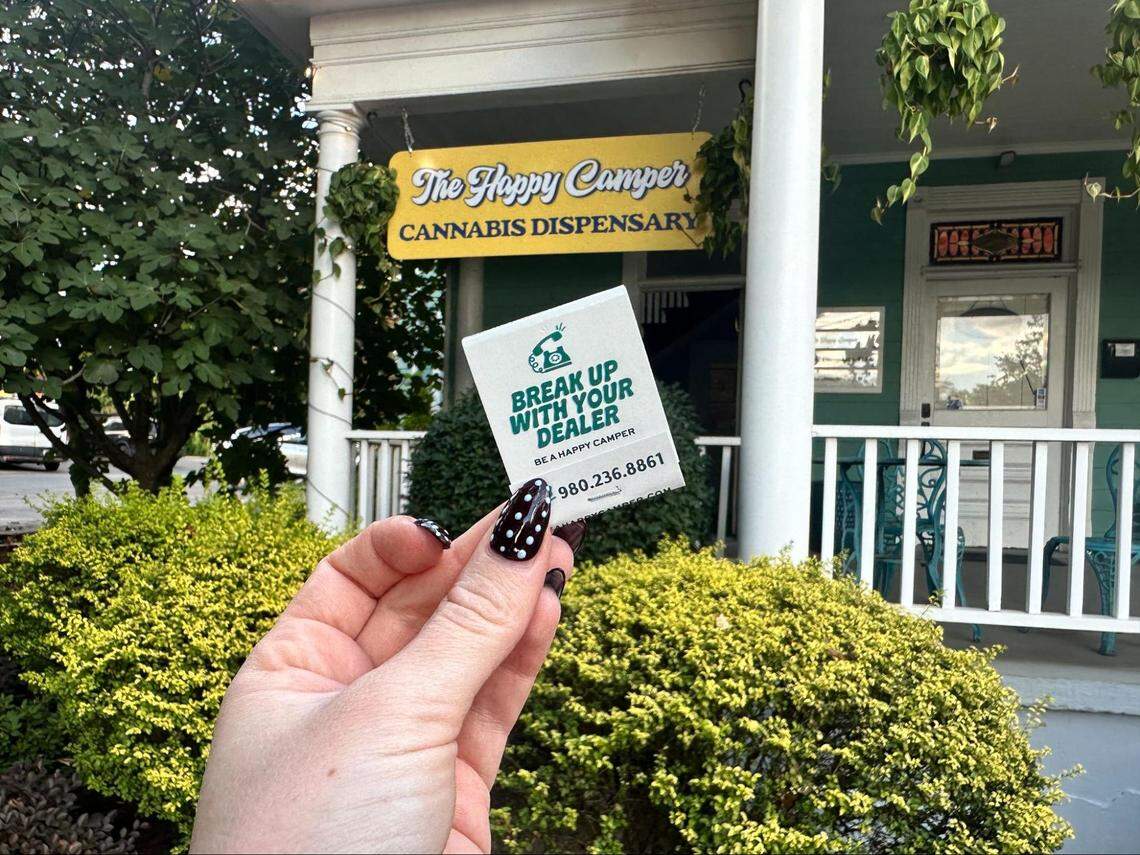 A person’s hand with black and white polka-dot nails holds up a matchbook with the text “BREAK UP WITH YOUR DEALER” in front of a storefront. The matchbook also says “BE A HAPPY CAMPER” and lists a phone number. In the background, a hanging yellow sign identifies the business as “The Happy Camper CANNABIS DISPENSARY.” The storefront has a porch and is surrounded by lush green bushes.