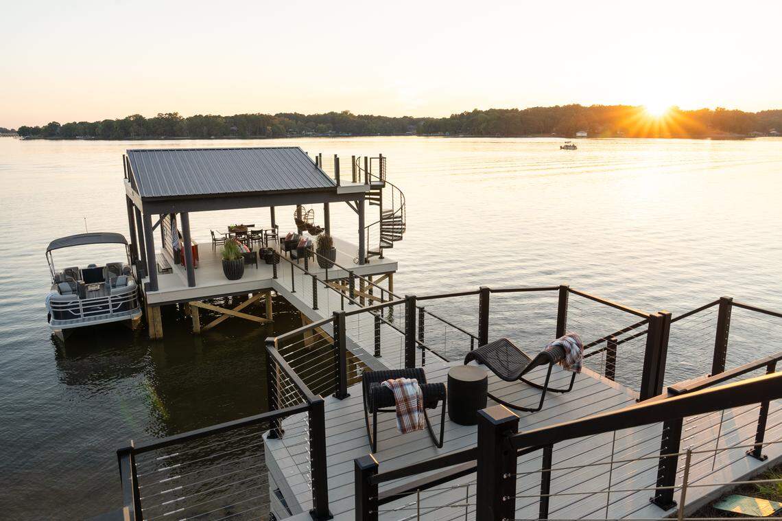 A multi-level, modern boat dock on a lake at sunset. The dock features a gray composite deck, black metal railings with thin cable wires, and a covered boat slip area with a dark metal roof. A black, spiral staircase leads to a small upper deck. A pontoon boat is tied to the side of the dock. In the foreground, the lower deck has two dark woven chaise lounges and a small round table. The sun is setting over the distant treeline across the calm water, casting a warm, golden glow across the scene.