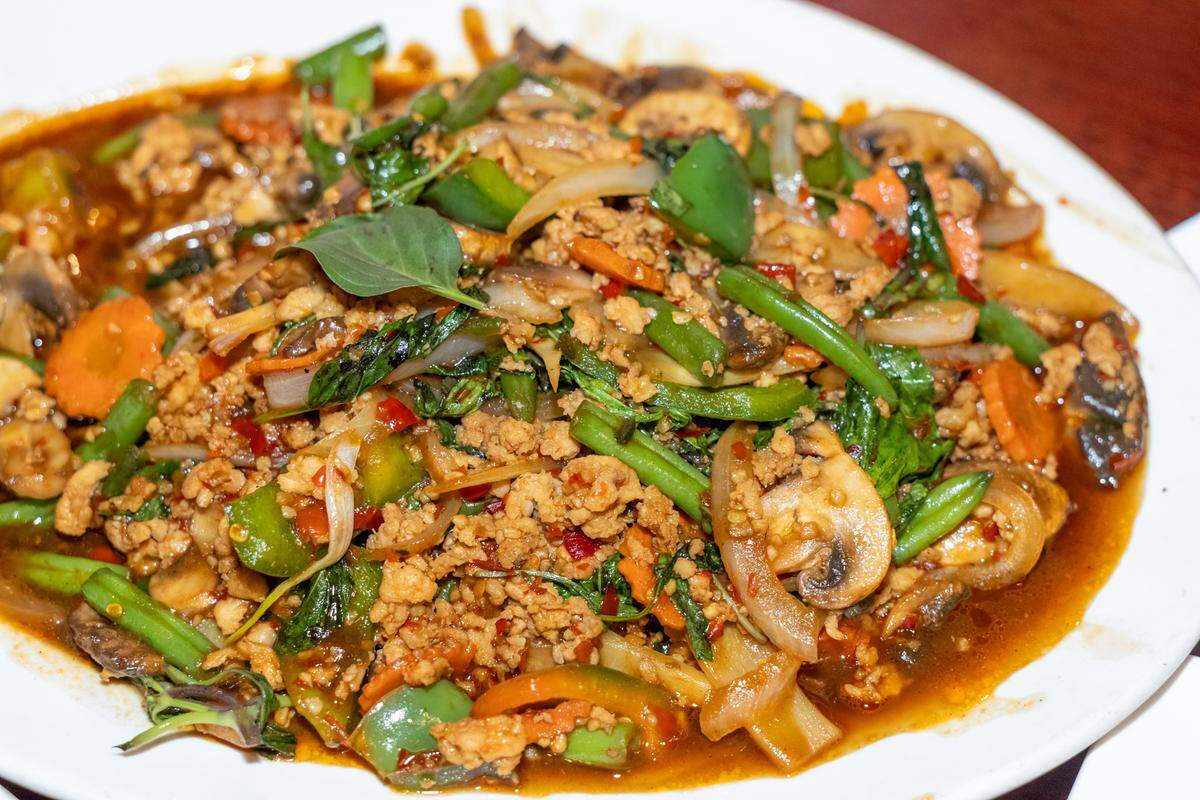 A close-up, top-down shot of a Thai stir-fry dish featuring minced meat, sliced mushrooms, green beans, carrots, and peppers in a savory brown sauce, garnished with fresh basil leaves.