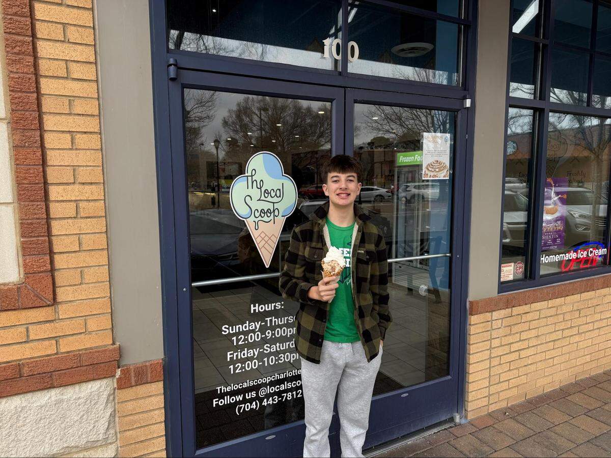 A smiling young person stands outside the front entrance of an ice cream shop holding a waffle cone with ice cream. They are wearing a green and black plaid jacket, a green t-shirt, and grey sweatpants. The entrance consists of glass double doors featuring a large logo for “The Local Scoop,” along with the store’s hours and phone number printed on the glass. The building exterior is made of tan brick.