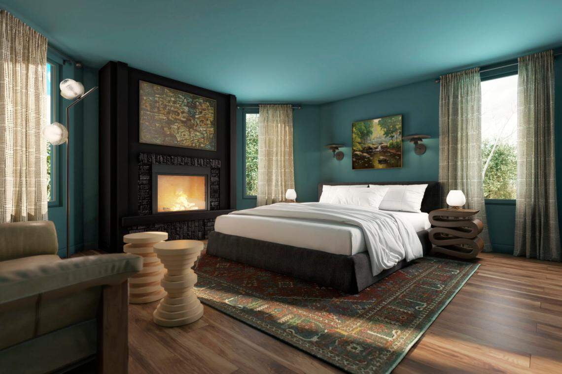 A cozy, eclectic hotel bedroom with teal blue walls and a matching ceiling. The room features a large bed with white bedding centered on a dark area rug. To the left, a large black fireplace mantel frames a roaring fire and a piece of abstract art. Two sets of stacked, light wood side tables sit near the fireplace. Large windows with patterned sheer curtains flank the room.