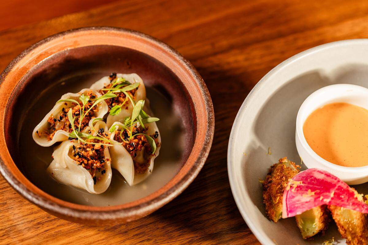 A high-angle close-up of two small plates on a wooden table. On the left, a rustic brown bowl contains three delicate steamed dumplings sitting in a light broth, topped with crunchy orange crumbles and fresh microgreens. On the right, a gray plate holds fried appetizers served with a small white bowl of creamy orange dipping sauce and a bright pink radish garnish.
