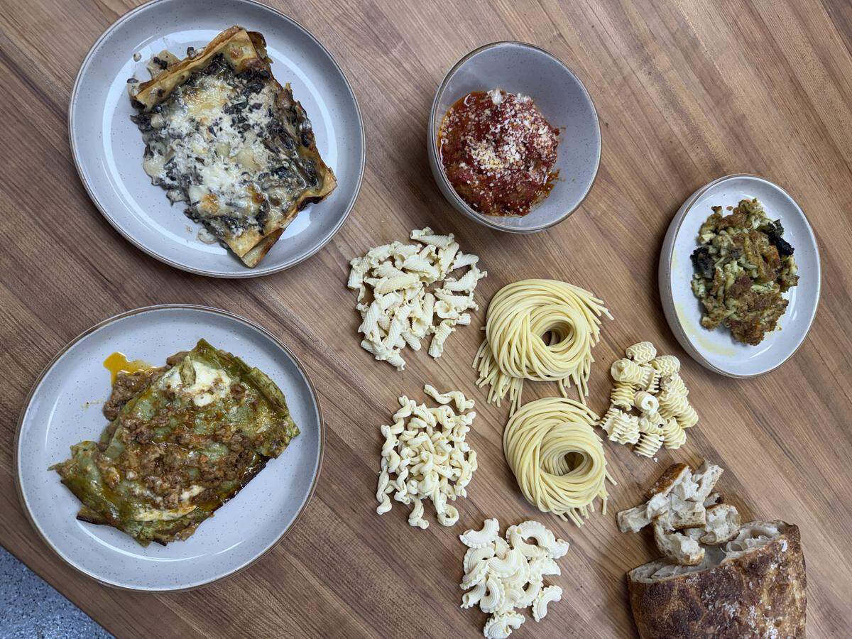 A top-down view of a wooden table featuring an array of fresh pasta shapes, a loaf of crusty bread, two portions of lasagna, a bowl of red sauce, and a side dish.
