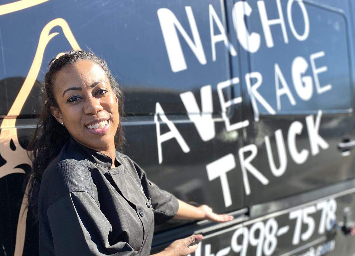 A smiling chef in a black chef’s coat stands in front of a black food truck. The chef is looking directly at the camera with a friendly expression and is leaning on the side of the truck, which has “NACHO AVERAGE TRUCK” and a phone number painted on it in large white letters. The sun is shining.