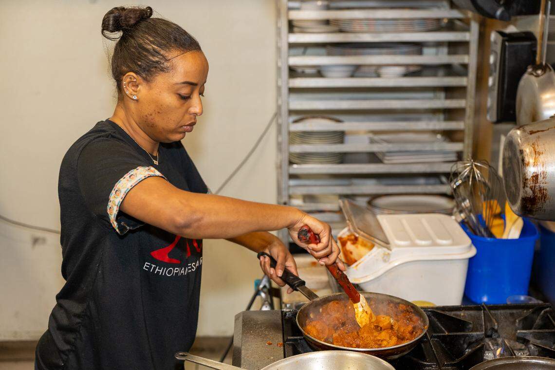A focused shot of a chef at the stove, using a spatula to stir a rich, red-hued stew in a pan while wearing a black shirt with “Ethiopian Restaurant” visible on the front. The kitchen is organized with stainless steel surfaces, a rack of clean plates, and various cooking utensils, showcasing the daily craft of Ethiopian cooking.