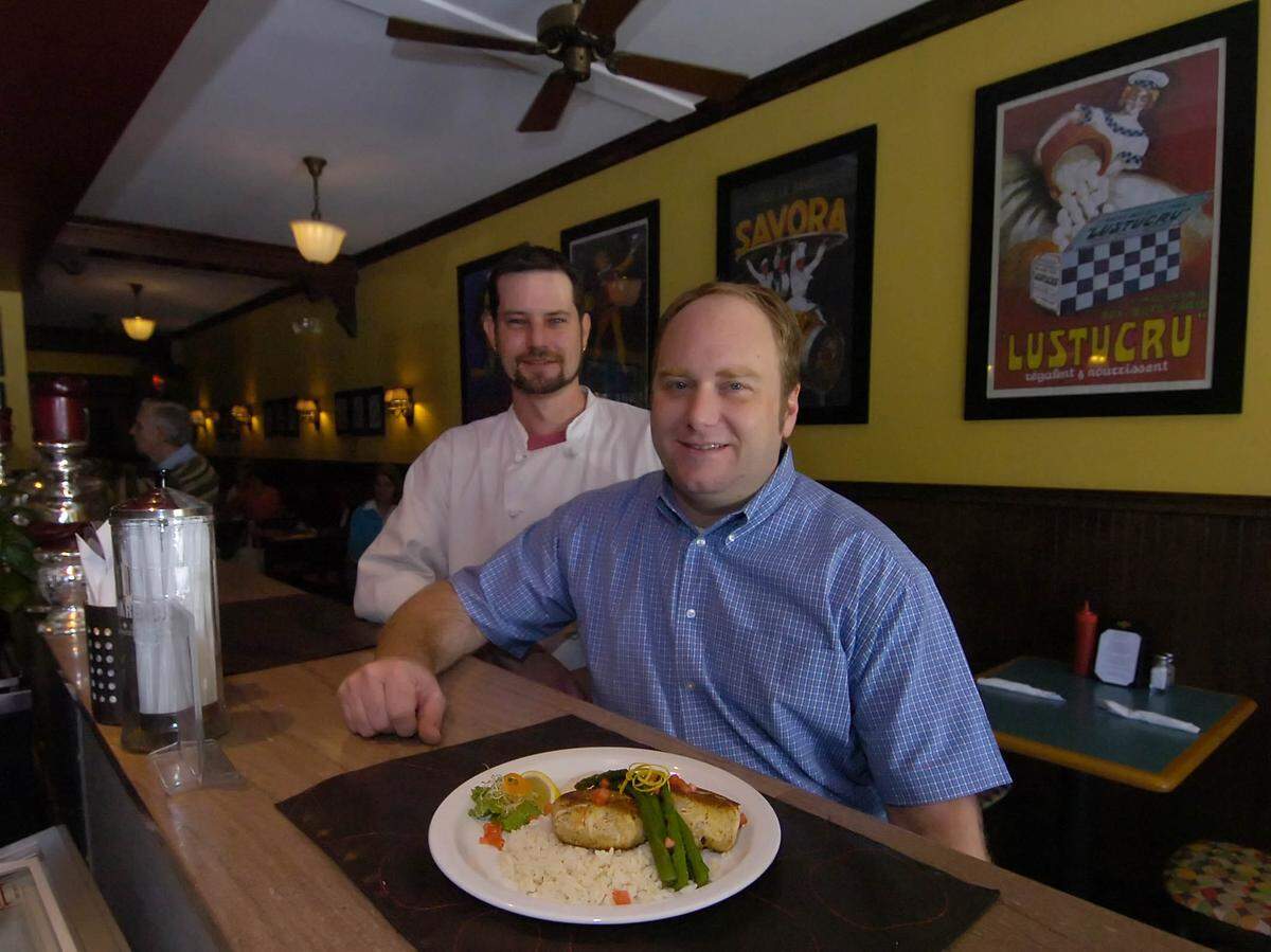 A 2007 photograph of two men—one in a blue checkered button-down and another in a white chef’s coat—smiling behind a dark wooden restaurant counter. In the foreground, a white plate is presented with a gourmet meal of crab cakes, white rice, and fresh asparagus. The background features warm yellow walls decorated with framed vintage French posters (”Lustucru” and “Savora”), dark wood trim, and a classic ceiling fan.