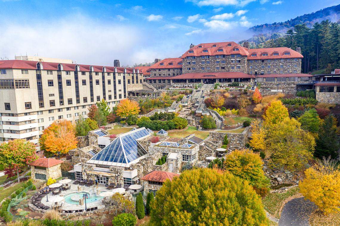 An elevated daytime view of the historic Omni Grove Park Inn in Asheville, North Carolina, during autumn. The grand stone hotel, with its iconic red roof, sits on a terraced hill surrounded by trees in peak yellow and orange fall foliage. In the foreground is the modern spa complex featuring a glass pyramid roof and outdoor pools.