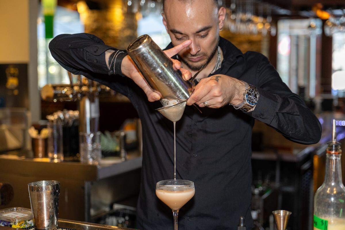 A focused shot of a bartender in a black shirt with a hand tattoo and wearing a silver watch carefully straining a peach-colored cocktail from a metal shaker into a coupe glass.
