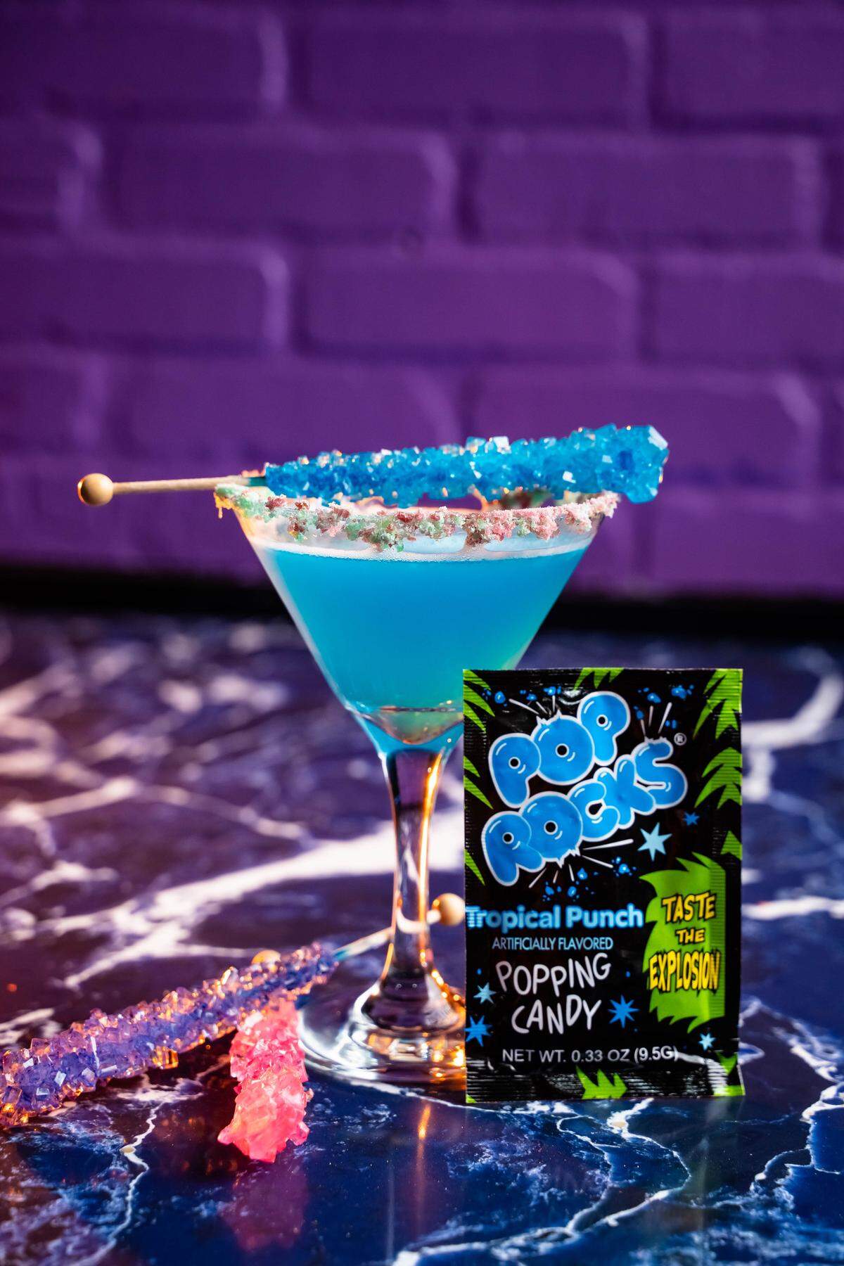 A bright blue cocktail in a martini glass is garnished with a blue rock candy stick and has a Pop Rocks-dusted rim. A packet of Tropical Punch Pop Rocks sits next to the glass.