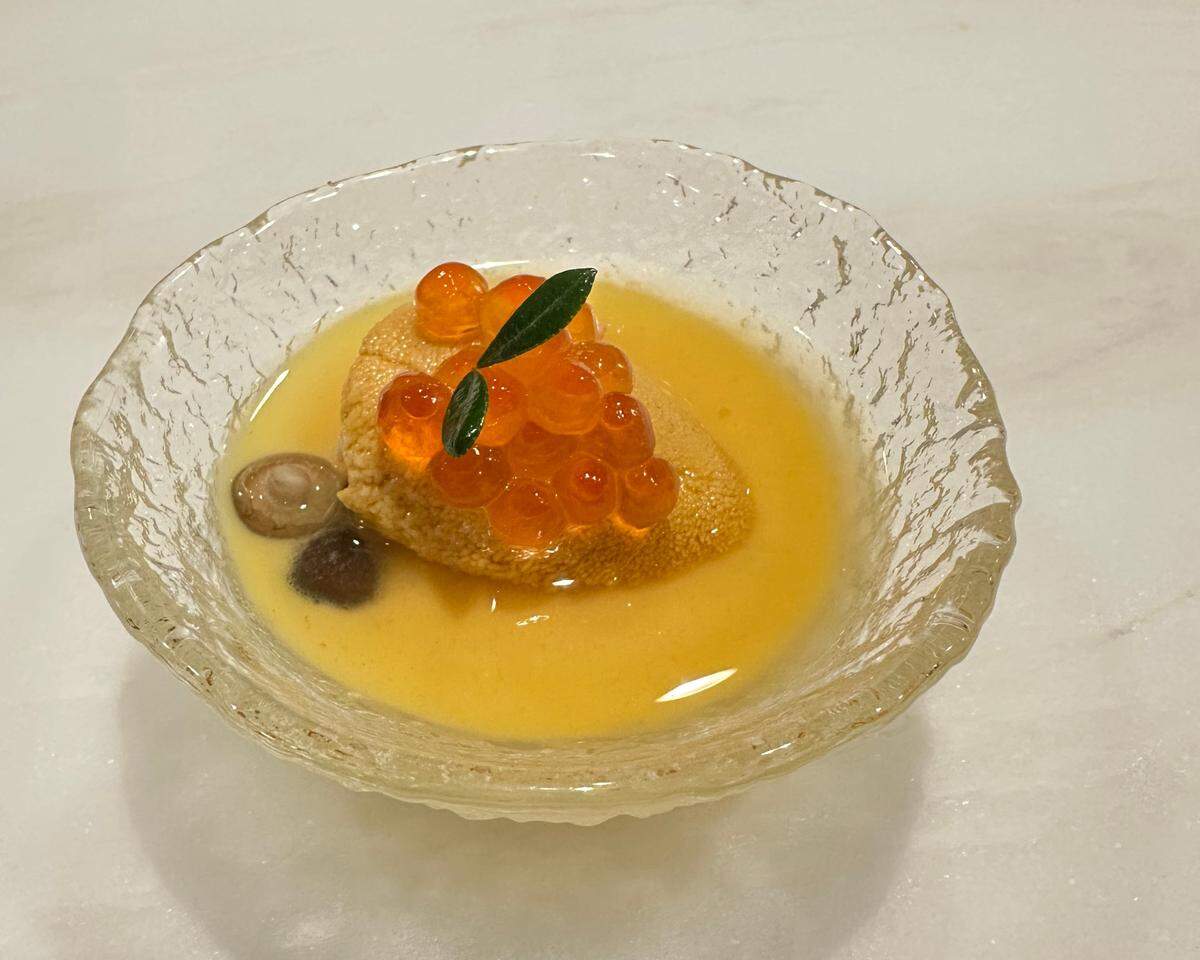 A sophisticated appetizer from Sora restaurant is presented in a textured glass bowl on a white surface. The dish features a lobe of uni and two small mushrooms sitting in a smooth yellow sauce. The uni is generously topped with large, translucent salmon roe and garnished with a sprig of a tiny green leaf.