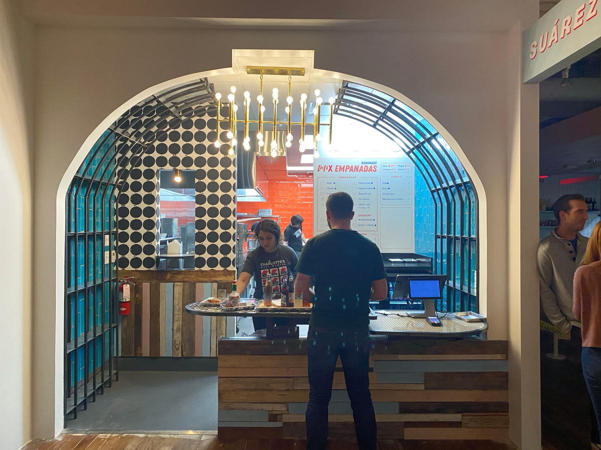A man in a dark green t-shirt stands at the wooden counter of Felix Empanadas, placing an order with a female employee. The scene is viewed through a large white archway, revealing a uniquely designed interior with a curved, teal metal cage structure, a wall with a bold black-and-white circular pattern, and a modern, multi-bulb chandelier hanging overhead.