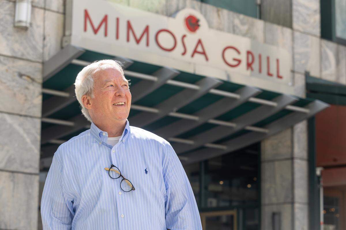 A portrait of person with white hair and a blue striped button-down shirt standing outside, smiling and looking upward, with the “Mimosa Grill” restaurant sign blurred in the background.