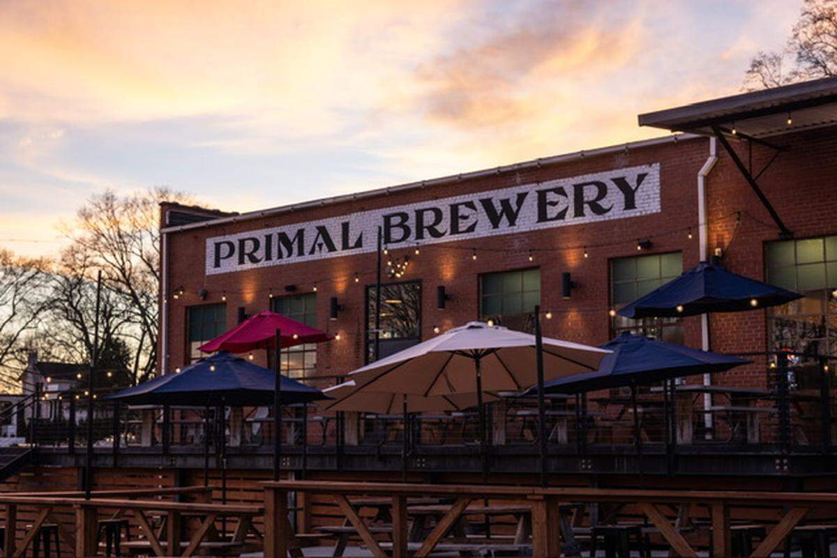 An exterior shot of the Primal Brewery building, a classic red brick structure with “PRIMAL BREWERY” painted in large white letters. The foreground features an outdoor patio with wooden picnic tables and large patio umbrellas under a soft, sunset sky.