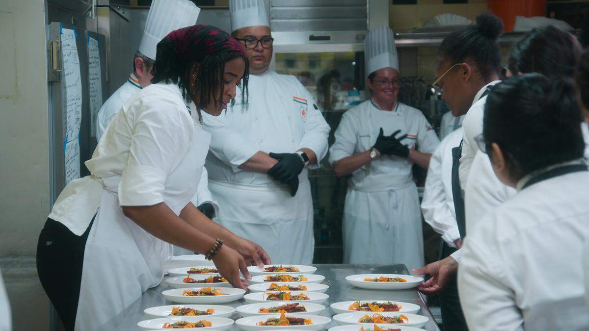 A dynamic scene in a professional kitchen where a team of chefs is plating dishes. The focus is on a chef with a red patterned headwrap who is intently adding finishing touches to a long line of identical, artistically presented dishes on white plates. Several other chefs, some in tall white toques, surround that chef, watching and assisting on the plating line.