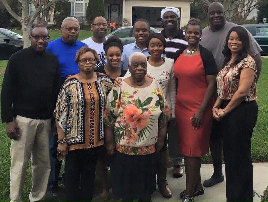 A multi-generational African American family of eleven people posing for a group photo outdoors in front of a suburban house. In the foreground, a woman is centered, smiling and wearing a white, black, and orange floral print top. She is flanked by two women in the middle row: one on the left wearing a colorful, patterned tunic and glasses, and one on the right in a white top with black floral accents. Behind them, there are several men and women in the back row, including one man in a bright blue shirt, another in a white T-shirt, and one in a striped shirt and bandana. The group is standing on a lawn, with a gray house and driveway visible in the background.