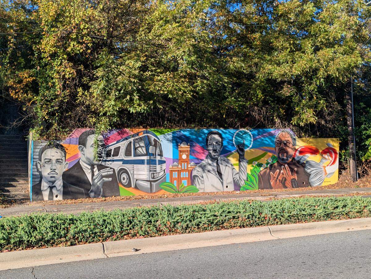 A wide, eye-level shot of a long, colorful street mural on a concrete retaining wall, set on a sunny day. The mural features realistic portraits of several African American men, including some in suits and one in a lab coat, alongside imagery like a white bus, all on a vibrant, abstract background. The wall is bordered by a low hedge and a road in the foreground, with dense green trees above.