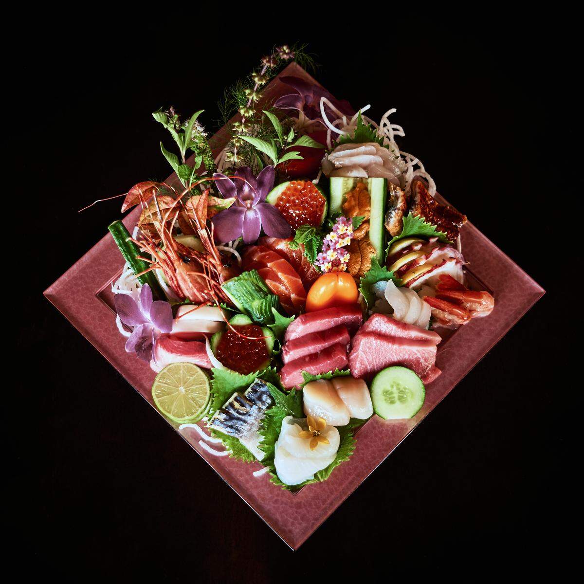 A vibrant and artfully arranged platter of sashimi and seafood on a reddish-pink, diamond-shaped plate, set against a black background. The platter is filled with various types of fresh fish, including red tuna and salmon, along with scallops, sea urchin, and shrimp with their heads still on. It is garnished with slices of lime and cucumber, purple orchids, fresh herbs, and a mound of bright orange fish roe.