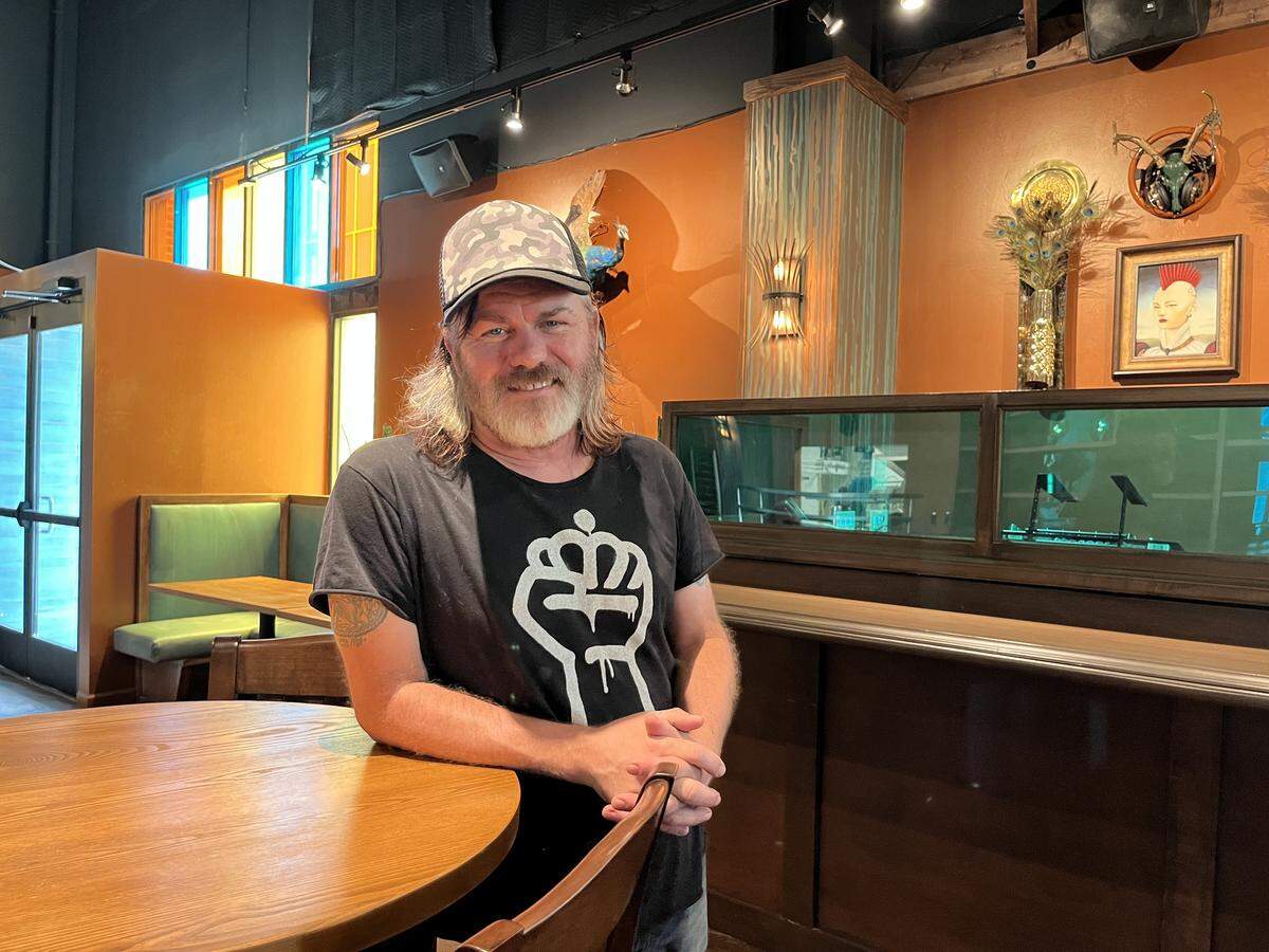 An interior designer with long light hair, a beard, and a camouflage cap leans against a round wooden table while wearing a black t-shirt with a white raised fist logo.