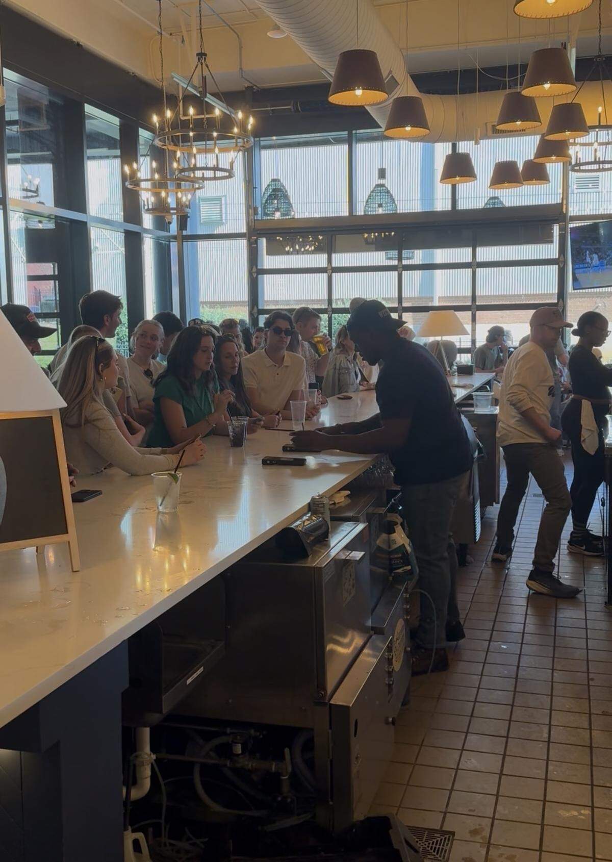 An indoor bar area with a long white countertop where patrons are gathered and talking. A bartender in a black shirt and baseball cap is standing behind the bar, interacting with customers. The space features modern lighting with multiple cone-shaped pendant lamps and circular chandeliers. Large windows and a glass garage door allow natural light to fill the room, showing a glimpse of buildings outside. The floor is tiled, and the overall atmosphere is busy and social.