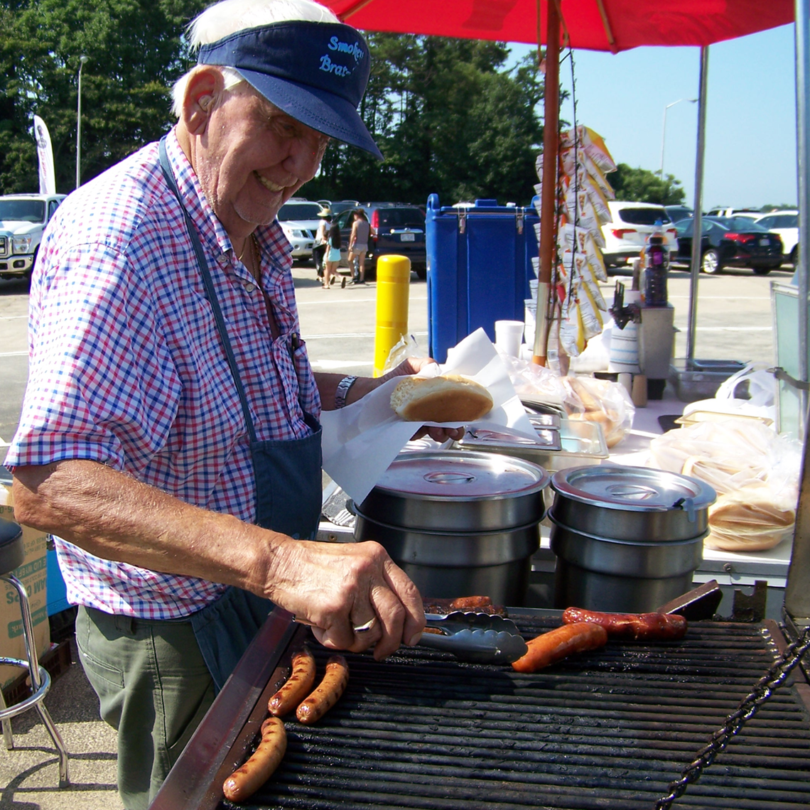 A food vendor stands behind a grill, cooking sausages and hot dogs. The vendor is wearing a blue visor that says “Smokin’ Brat,” a red and blue checkered short-sleeve shirt, and a blue apron. The individual is smiling and using a pair of tongs to flip a sausage on the grill in one hand, while in other hand is a hot dog bun wrapped in a small sheet of paper. The food cart is set up with stainless steel warming pots and a red umbrella for shade. In the background, a parking lot with cars and people is visible.