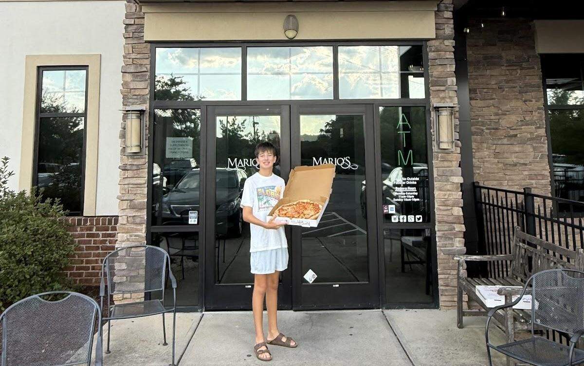 A smiling teenage boy with brown hair, wearing a white t-shirt and light-colored shorts, stands outside the entrance of “Mario’s” restaurant. He is holding a large, open pizza box containing a round cheese pizza. The restaurant’s storefront is made of glass doors and stone, with “MARIO’S” written in script on the windows. A sign with business hours and social media logos is visible on the right door. There are several outdoor tables and chairs in the foreground, and the reflection of clouds and a street are visible in the glass doors.