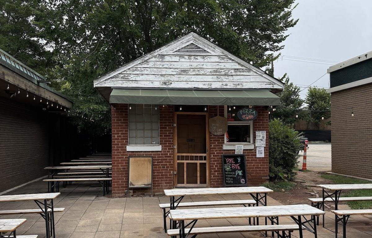 A wide shot of a pizzeria, which operates out of a small, standalone brick building with a screen door and a takeout window. In front of the building is a spacious patio with rows of light-colored wooden picnic tables. The scene is set under an overcast sky with trees in the background.