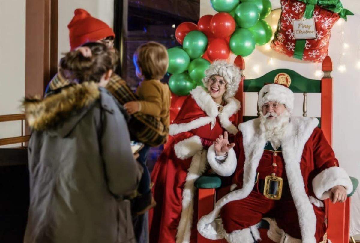 A Santa performer and Mrs. Claus, both in traditional red and white velvet attire, sit in a large wooden chair decorated with a holiday wreath. They are smiling and waving at a family with a small child standing in the foreground, set against a festive backdrop of red and green balloons.