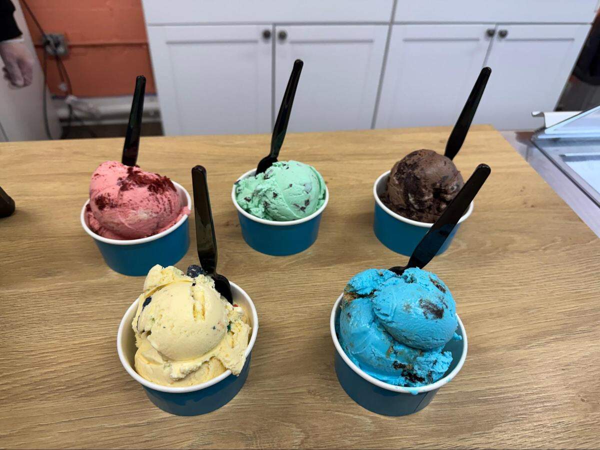 A high-angle shot of five different flavors of ice cream in small blue cups arranged on a wooden counter. The flavors include strawberry, mint chocolate chip, chocolate brownie, cake batter with sprinkles, and a bright blue “cookie monster” style flavor. Each cup has a black plastic spoon.