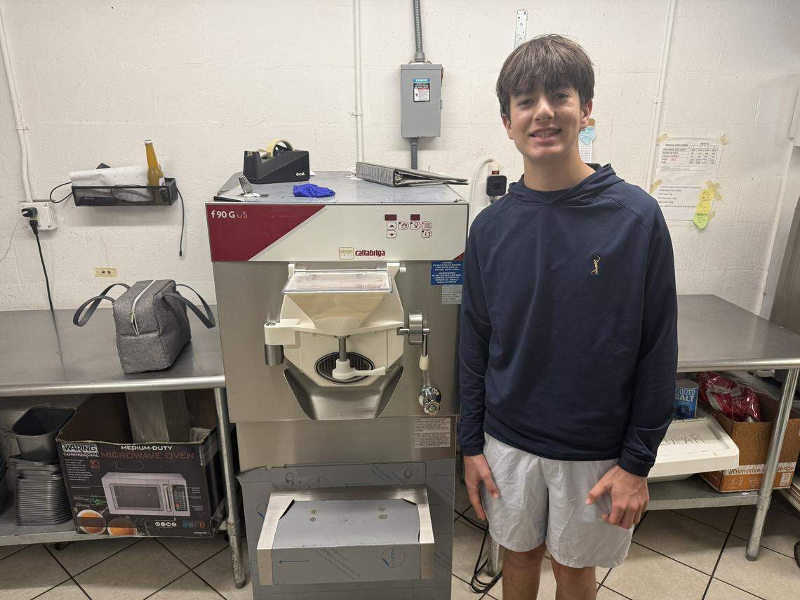 A young person in a navy hoodie and grey shorts stands in a professional kitchen area next to a large, stainless steel Cattabriga gelato machine. The industrial setting features white brick walls and stainless steel prep tables, giving a glimpse into where the handcrafted desserts are produced.