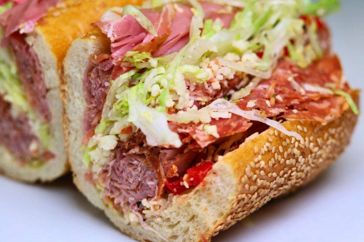 An extreme close-up of an Italian hoagie on a sesame seed roll. The sandwich is cut, showcasing its generous fillings of various cured meats, including salami and capicola, topped with shredded lettuce and grated cheese.
