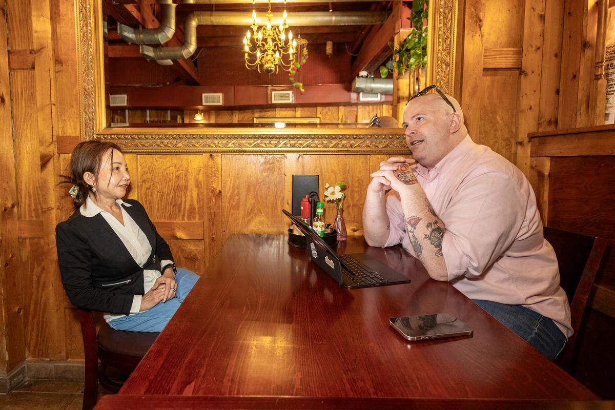 A restarateur in a black blazer and a writer with arm tattoos in a pink button-down sit across from each other at a wooden restaurant table, engaged in conversation. A laptop and a smartphone are on the table between them in a cozy, wood-paneled room.