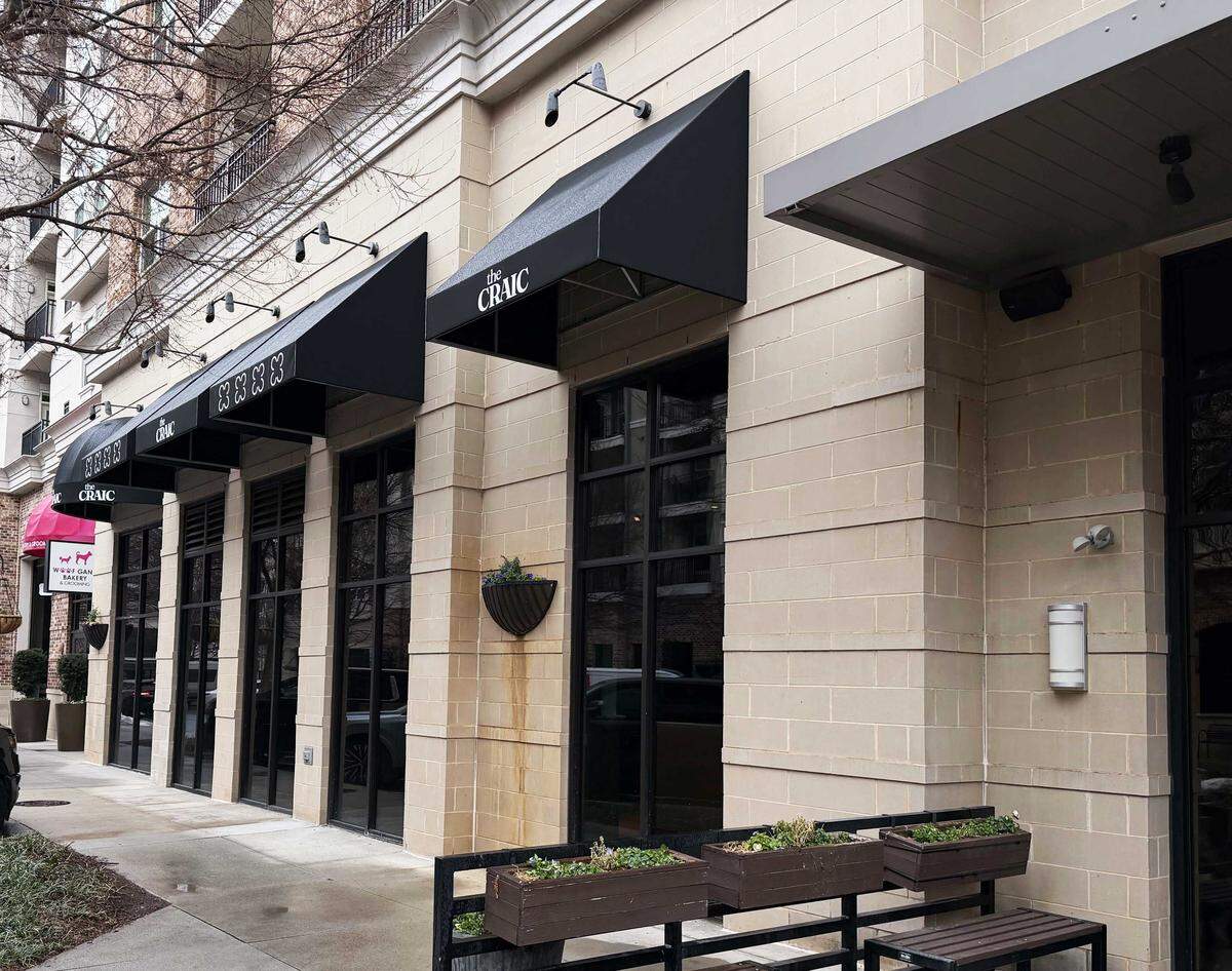 The exterior of the restaurant shows a series of large black-framed windows set into a light tan stone building under several black awnings. The awnings are branded with “the CRAIC” and a four-leaf clover motif, overlooking a sidewalk with wooden planters.