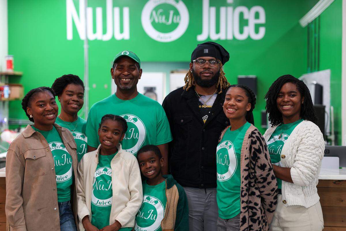 A smiling group of eight people, appearing to be a family, posing for a photo inside a shop. They are standing in two rows in front of a bright green wall featuring the large white logo “NuJu Juice”. Most of the family members are wearing matching green t-shirts with the business logo.