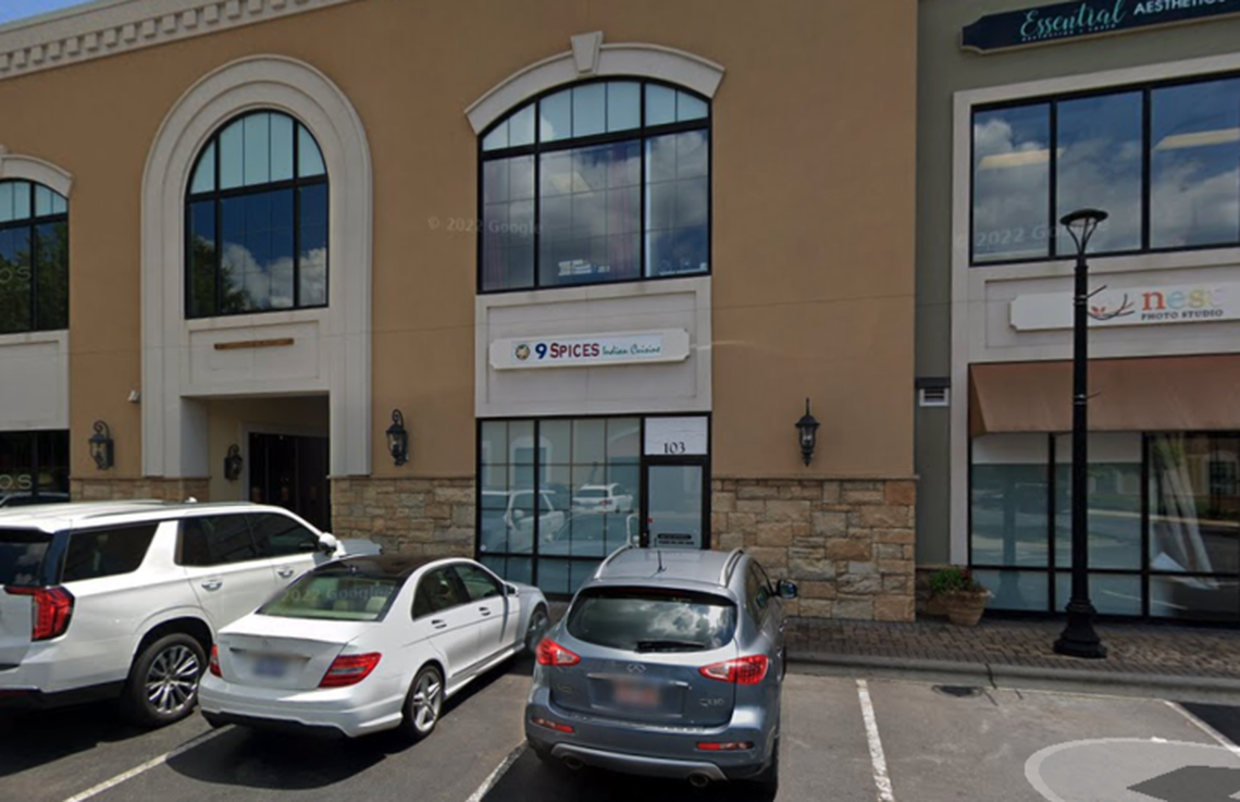 A slightly angled shot of a tan and stone-faced strip mall building. The storefront in the center has a large arched window on the second floor and a sign for “9 SPICES Indian Cuisine” over the entrance. A small plant is visible in a pot near the door. Several cars are parked in the lot in front, including a gray SUV and a white sedan. Another storefront with a sign for “Essential Aesthetics” is visible on the right.