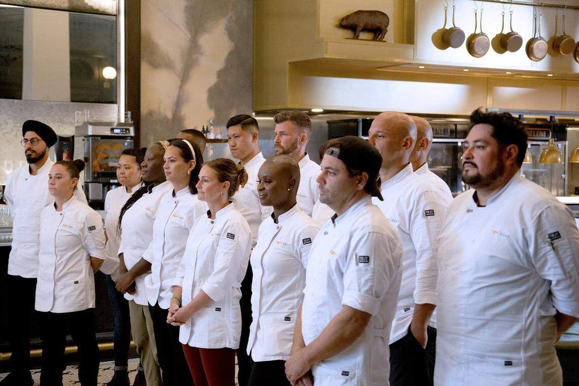 A group of chefs in white professional chef coats stand in a restaurant kitchen. The lighting is warm, and copper pots hang from a rack in the background.