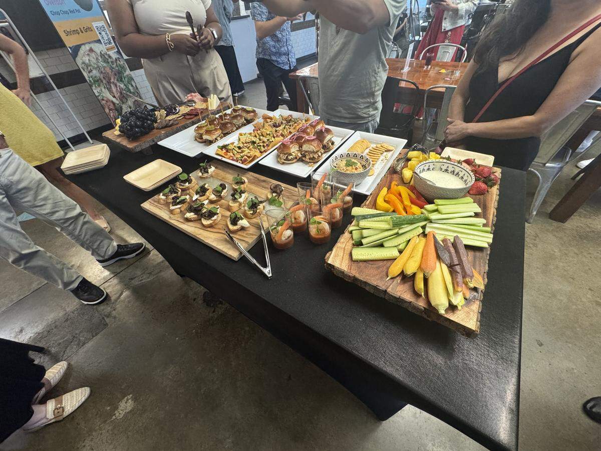 A high-angle, wide shot of an extensive buffet spread arranged on a long table covered with a black tablecloth. The food is displayed on various platters and wooden boards, including a large crudité platter with sliced cucumbers, bell peppers, celery, and a dipping bowl. Other items feature small slider sandwiches, an assortment of bruschetta on a wooden board, small plastic cups containing shrimp cocktail, and a charcuterie board with grapes and crackers. Several people are gathered around the table, partially visible from the waist down or side profile, waiting to serve themselves.