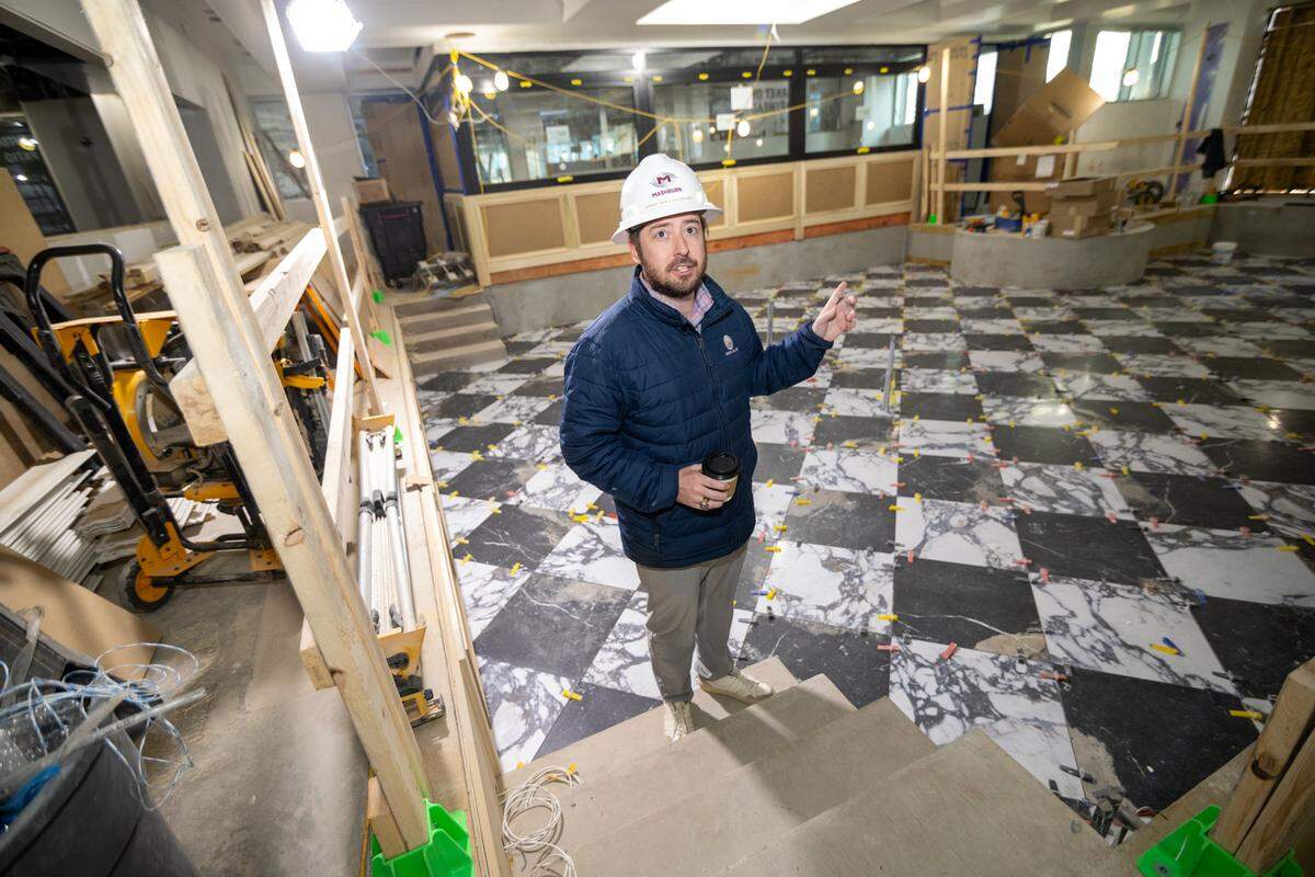 A construction professional wearing a white hard hat and blue puffer jacket stands on concrete steps, gesturing toward the interior of the checkered-tiled room while holding a coffee cup.